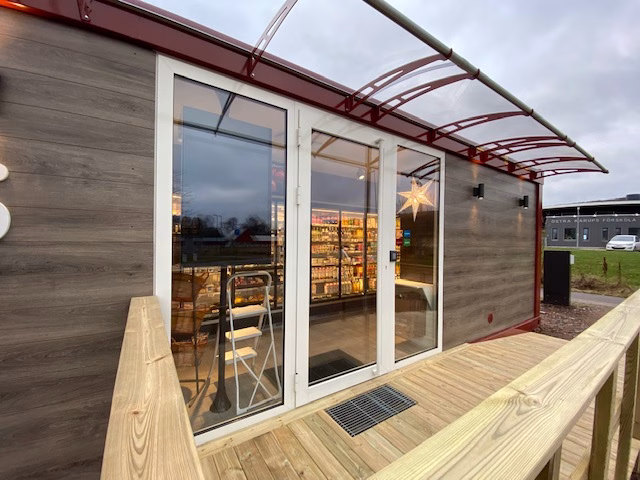 A modern prefabricated shop with a wood-paneled exterior, featuring double glass doors leading to a brightly lit interior, a wooden deck, and a clear awning.